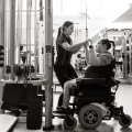 A young women seated in a wheelchair is working at a pull-down weights machine in a gym, assisted by a female coach. The photo is in black and white.